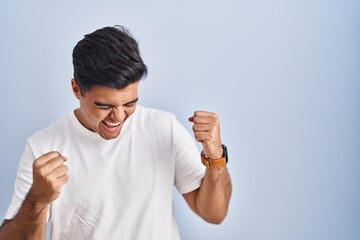 Hispanic man standing over blue background very happy and excited doing winner gesture with arms raised, smiling and screaming for success. celebration concept.