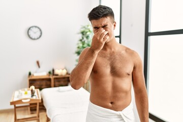 Young hispanic man standing shirtless at spa center smelling something stinky and disgusting, intolerable smell, holding breath with fingers on nose. bad smell