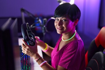 Middle age chinese woman streamer smiling confident holding headphones at gaming room