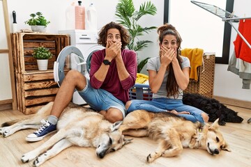 Young hispanic couple doing laundry with dogs shocked covering mouth with hands for mistake. secret...