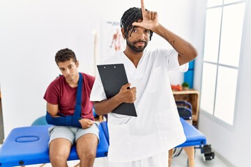Young hispanic man working at pain recovery clinic with a man with broken arm making fun of people with fingers on forehead doing loser gesture mocking and insulting. - Powered by Adobe