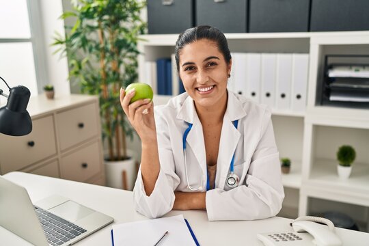 Young hispanic woman wearing dietitian uniform holding apple at clinic