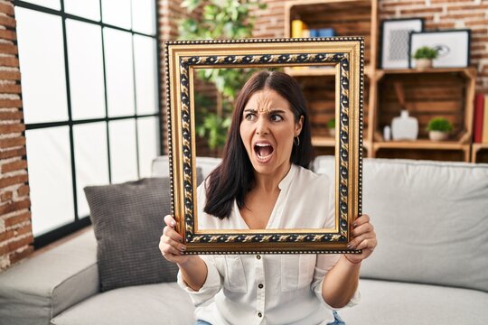 Young hispanic woman holding empty frame angry and mad screaming frustrated and furious, shouting with anger. rage and aggressive concept.