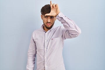 Arab man with beard standing over blue background making fun of people with fingers on forehead...