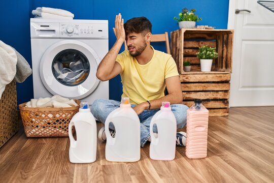 Arab man with beard doing laundry sitting on the floor with detergent bottle surprised with hand on head for mistake, remember error. forgot, bad memory concept.
