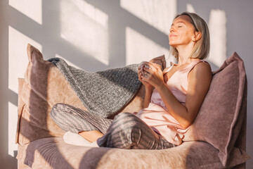 Happy dreamy middle aged woman sitting on sofa in living room with cup of tea or coffee - 534795102