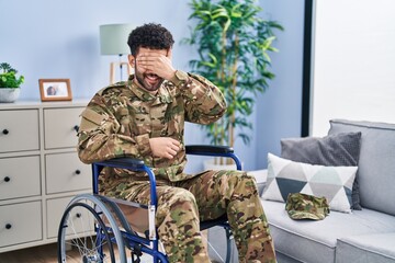 Fototapeta premium Arab man wearing camouflage army uniform sitting on wheelchair smiling and laughing with hand on face covering eyes for surprise. blind concept.