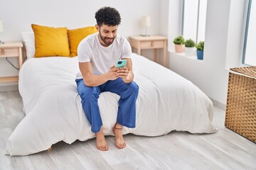 Young arab man using smartphone sitting on bed at bedroom