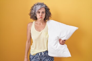 Middle age woman with grey hair wearing pijama hugging pillow puffing cheeks with funny face. mouth...