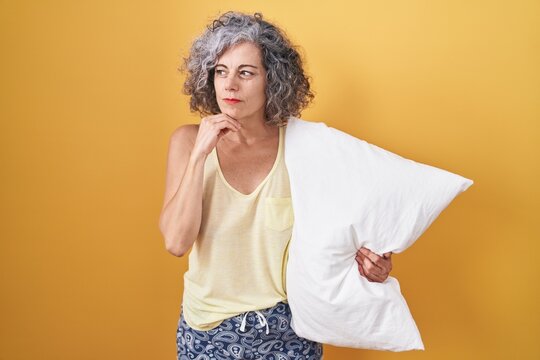 Middle age woman with grey hair wearing pijama hugging pillow with hand on chin thinking about question, pensive expression. smiling with thoughtful face. doubt concept.