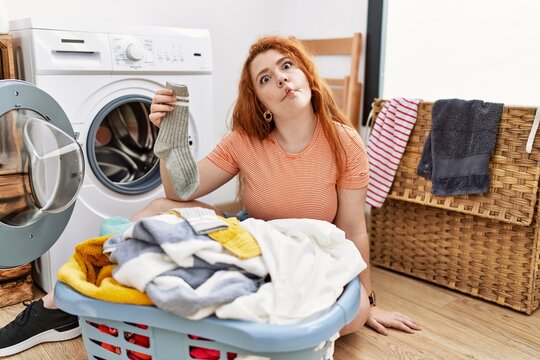 Young redhead woman putting dirty laundry into washing machine making fish face with lips, crazy and comical gesture. funny expression.