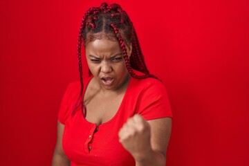 African american woman with braided hair standing over red background angry and mad raising fist...