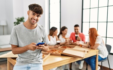 Group of young people eating italian pizza sitting on the table. Man smiling happy using smartphone at home.