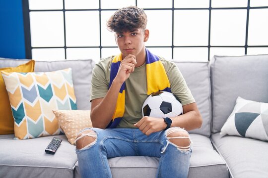 Hispanic teenager sitting on the sofa watching football match serious face thinking about question with hand on chin, thoughtful about confusing idea