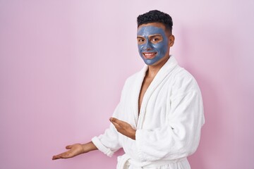 Young hispanic man wearing beauty face mask and bath robe inviting to enter smiling natural with open hand