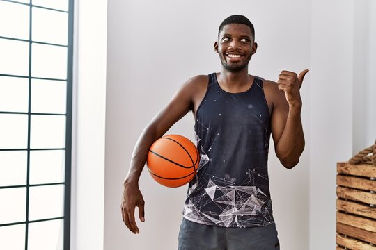 Young african american man holding basketball ball at home pointing thumb up to the side smiling happy with open mouth