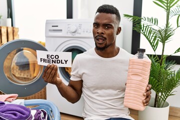 Young african man doing laundry holding eco friendly paper clueless and confused expression. doubt...