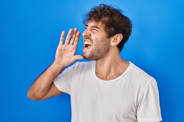 Hispanic young man standing over blue background shouting and screaming loud to side with hand on mouth. communication concept.