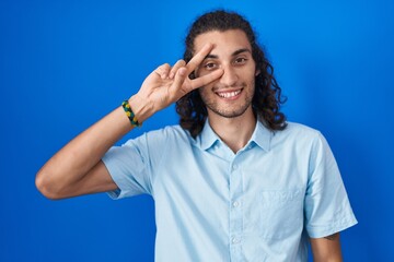 Young hispanic man standing over blue background doing peace symbol with fingers over face, smiling cheerful showing victory