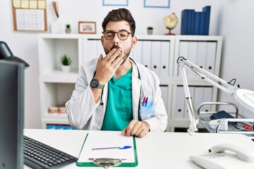 Young man with beard wearing doctor uniform and stethoscope at the clinic bored yawning tired covering mouth with hand. restless and sleepiness.