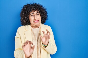 Young brunette woman with curly hair standing over blue background disgusted expression, displeased...