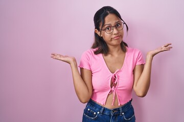 Hispanic young woman standing over pink background wearing glasses clueless and confused expression...