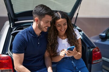 Young hispanic couple sitting on car trunk using smartphone at street