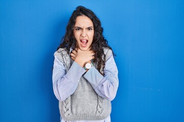 Obraz premium Young brunette woman standing over blue background shouting suffocate because painful strangle. health problem. asphyxiate and suicide concept.