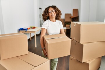 Middle age hispanic woman smiling confident holding cardboard box at new home