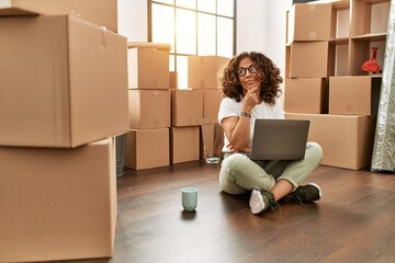 Middle age hispanic woman sitting on the floor at new home with laptop serious face thinking about question with hand on chin, thoughtful about confusing idea