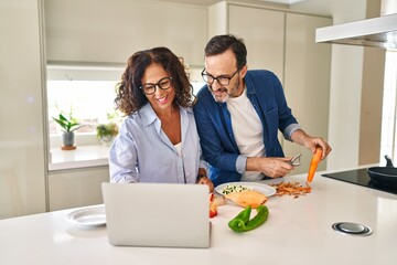 Middle age hispanic couple cooking and using laptop at kitchen
