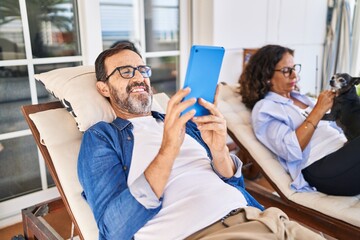 Middle age hispanic couple using touchpad lying on hammock with dog at terrace