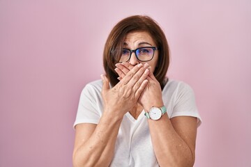 Middle age hispanic woman standing over pink background shocked covering mouth with hands for...