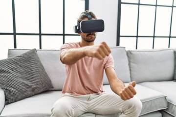 Handsome hispanic man wearing virtual reality glasses driving on videogame at the living room at home