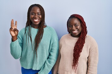 Two african woman standing over blue background showing and pointing up with fingers number three while smiling confident and happy.