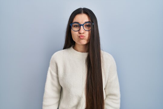 Young hispanic woman wearing casual sweater over blue background puffing cheeks with funny face. mouth inflated with air, crazy expression.