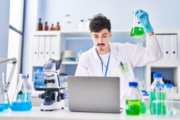 Young caucasian man scientist using laptop holding test tube at laboratory