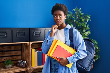 African american woman wearing student backpack and holding books serious face thinking about question with hand on chin, thoughtful about confusing idea