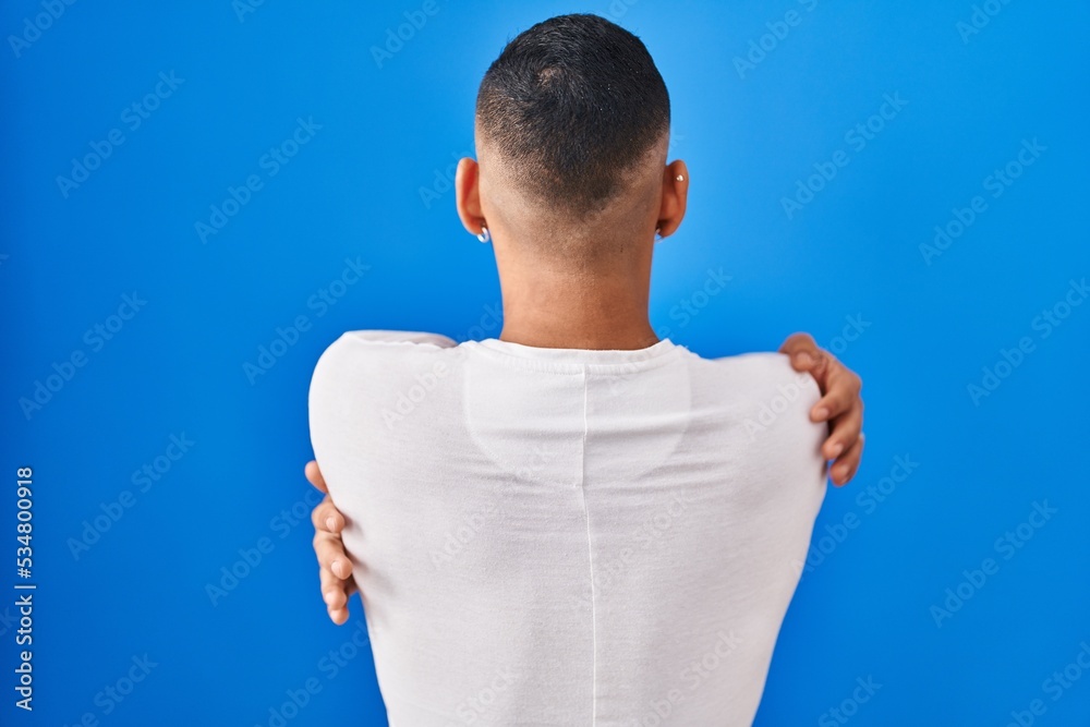 Canvas Prints Young hispanic man standing over blue background hugging oneself happy and positive from backwards. self love and self care