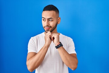 Young hispanic man standing over blue background laughing nervous and excited with hands on chin looking to the side