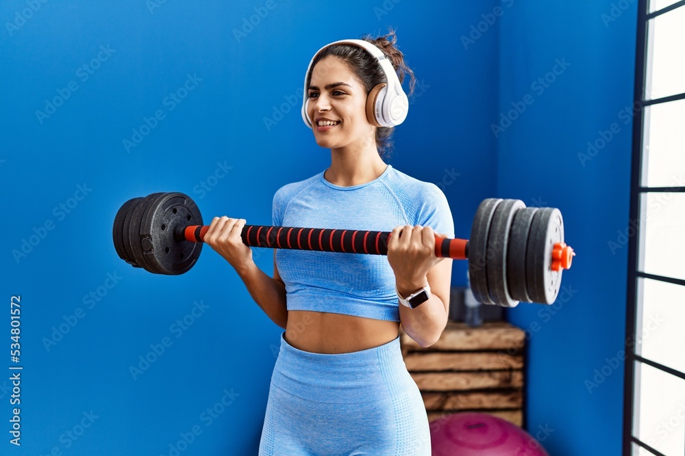 Wall mural young latin woman listening to music training using barbell at sport center