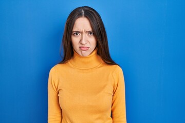 Young brunette woman standing over blue background skeptic and nervous, frowning upset because of...
