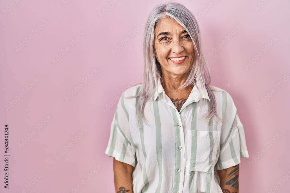 Wall mural middle age woman with grey hair standing over pink background looking positive and happy standing an