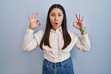 Young latin woman standing over blue background looking surprised and shocked doing ok approval symbol with fingers. crazy expression