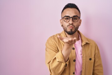 Fototapeta premium Young hispanic man standing over pink background looking at the camera blowing a kiss with hand on air being lovely and sexy. love expression.