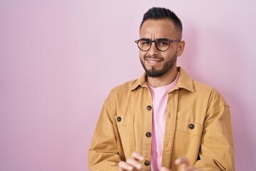 Young hispanic man standing over pink background disgusted expression, displeased and fearful doing disgust face because aversion reaction. with hands raised