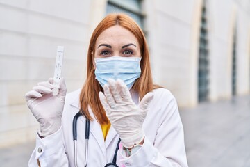 Young doctor woman holding coronavirus infection test covering mouth with hand, shocked and afraid...
