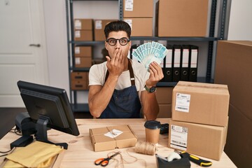 Young hispanic man working at small business ecommerce holding brazilian reals covering mouth with hand, shocked and afraid for mistake. surprised expression