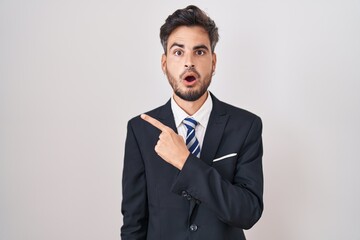 Young hispanic man with tattoos wearing business suit and tie surprised pointing with finger to the side, open mouth amazed expression.