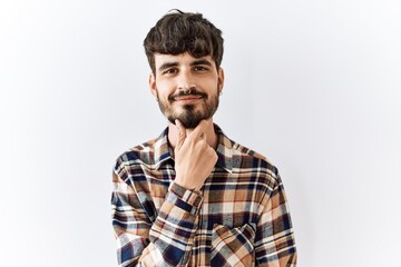 Hispanic man with beard standing over isolated background looking confident at the camera with smile with crossed arms and hand raised on chin. thinking positive.